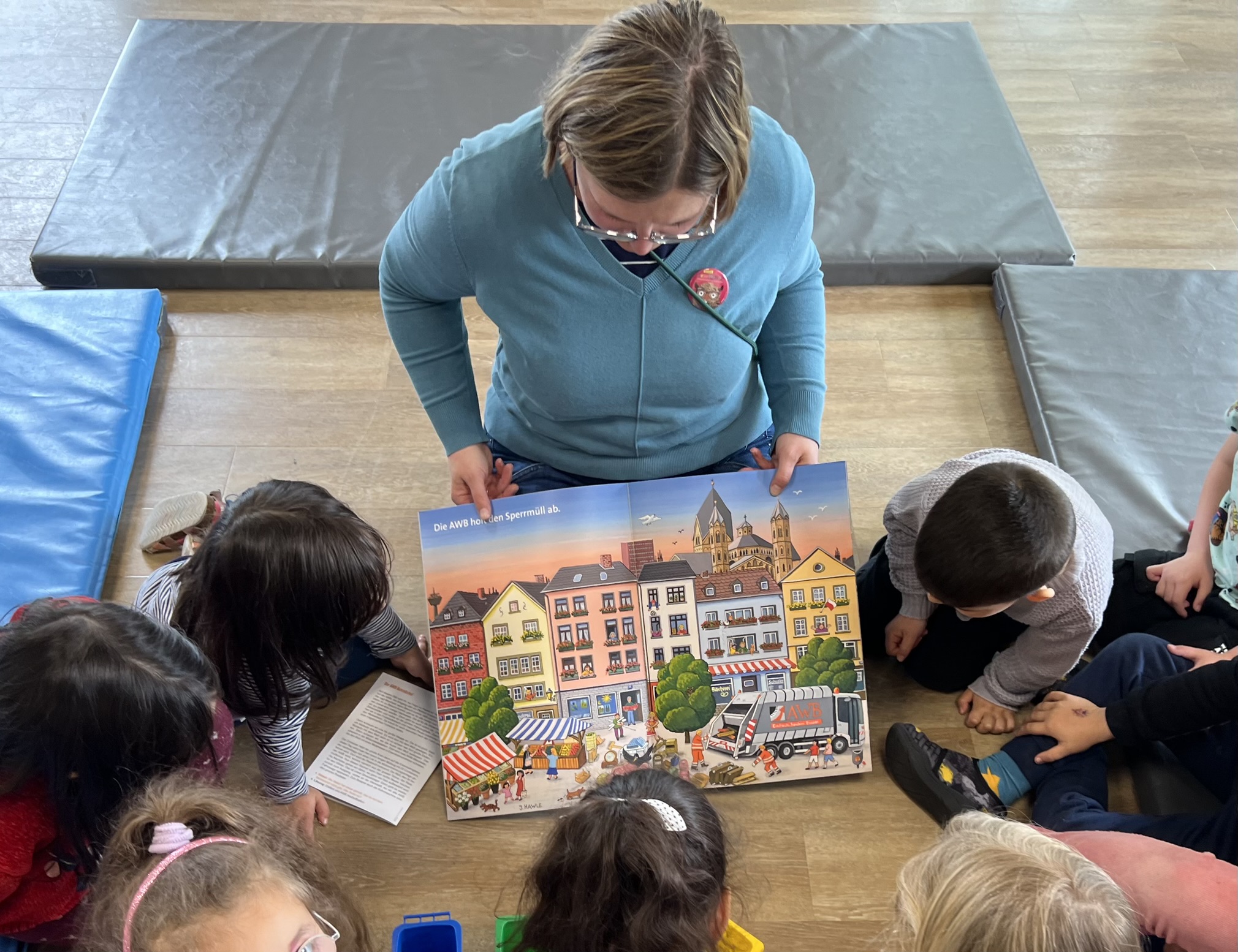 Vorleserin Anita Rupp sitzt mit einem geöffneten Bilderbuch vor Kindern. Die Kinder betrachten das Buch.