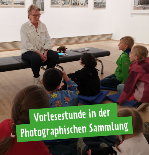 Eine Frau mit grauen kurzen Haaren in weißer Bluse sitzt auf einer Bank. Im Hintergrund hängen Bilder. Mehrere Kinder sitzen vor der Bank und sehen die Frau an.