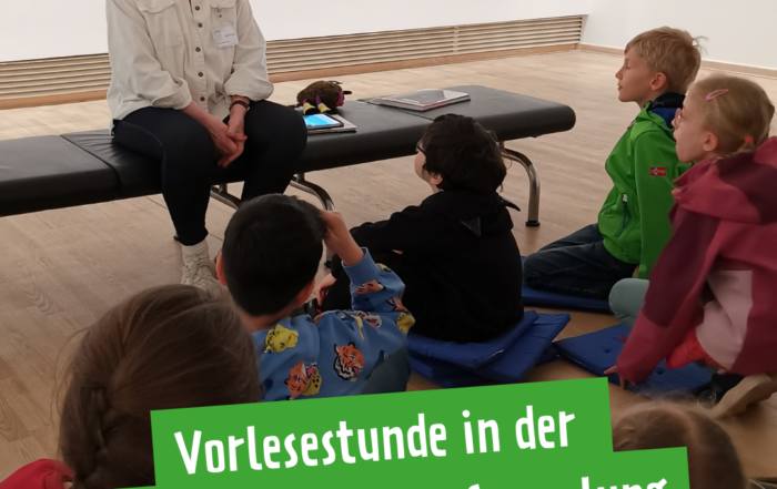 Eine Frau mit grauen kurzen Haaren in weißer Bluse sitzt auf einer Bank. Im Hintergrund hängen Bilder. Mehrere Kinder sitzen vor der Bank und sehen die Frau an.