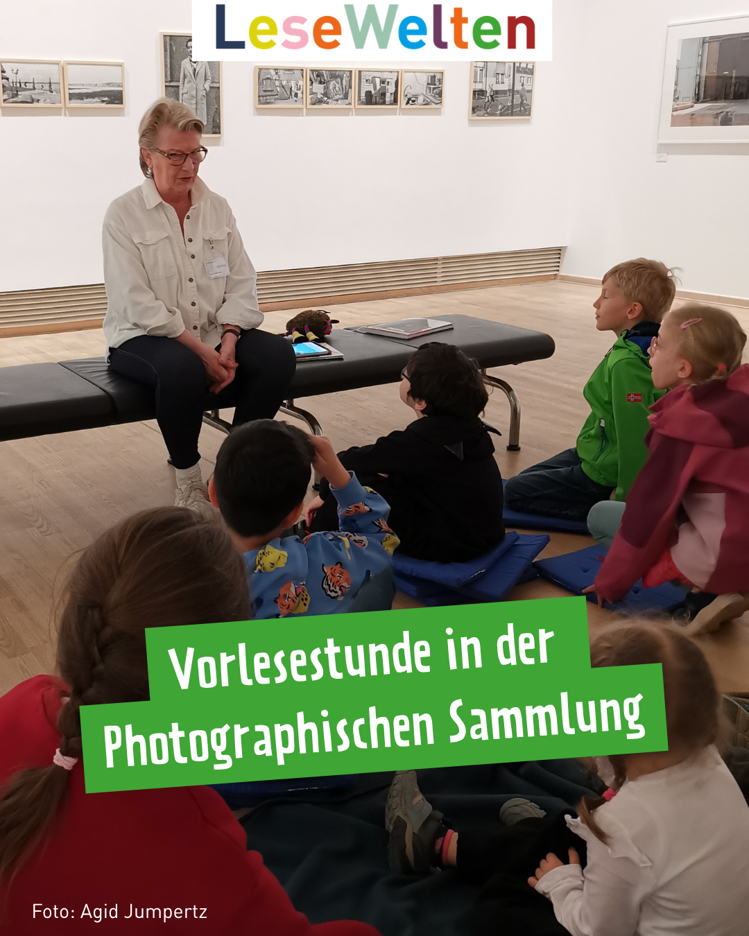 Eine Frau mit grauen kurzen Haaren in weißer Bluse sitzt auf einer Bank. Im Hintergrund hängen Bilder. Mehrere Kinder sitzen vor der Bank und sehen die Frau an.