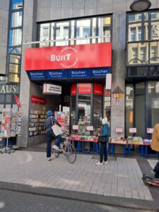 Ein Mann auf einem Fahrrad und eine Frau gehen an einer Buchhandlung mit der Aufschrift "Bunt - modernes Antiquariat" vorbei. Die Schaufenster sind voller Bücher.