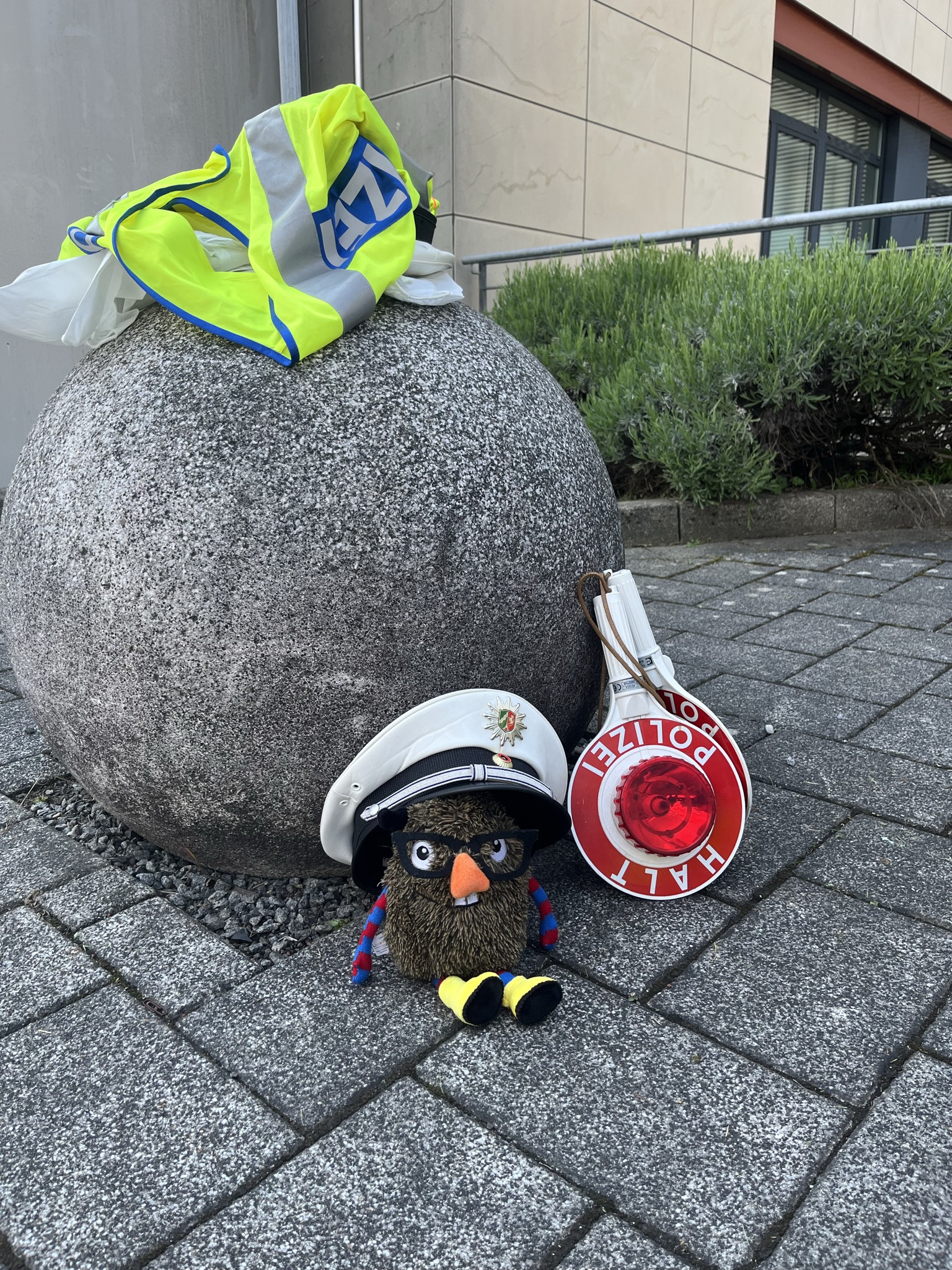 IMG_1137 Ein kleines Monster aus Stoff mit großer orangefarbener Nase und dicker Brille sitzt vor einem Stein. Es trägt eine Polizeimütze. Daneben liegt eine Polizeikelle und auf dem Stein eine Warnweste der Polizei.